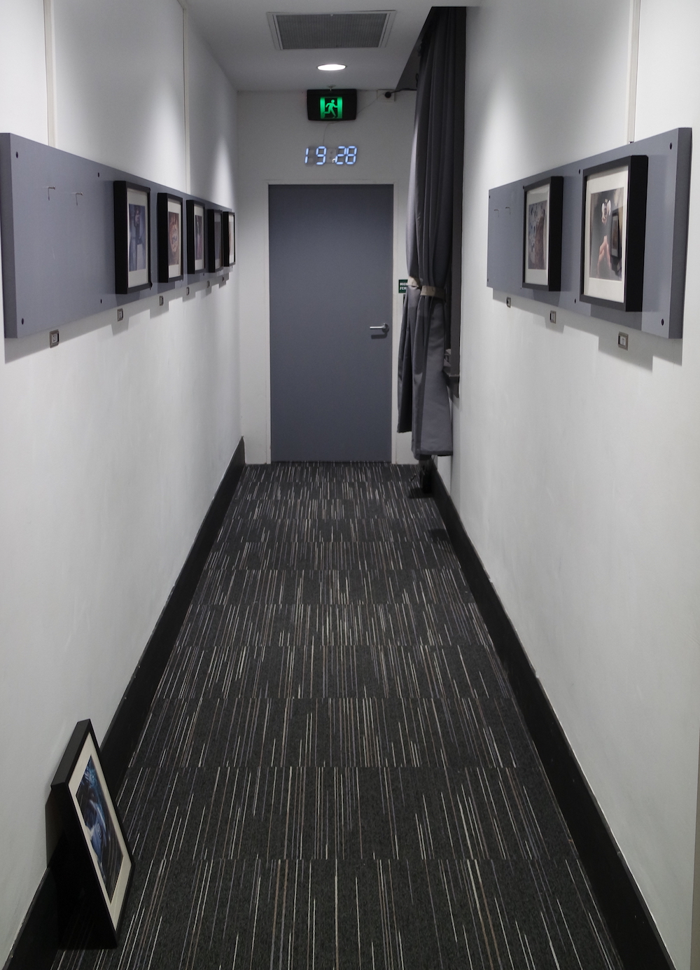 A long hallway with pictures mounted on the walls, an a picture on the floor leaned against the wall. The opposite wall has a door with a clock over it.
