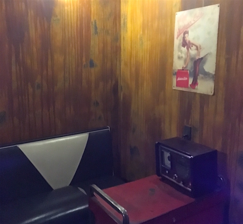 A view of the Vault. There is an old diner bench, a workbench, and a large radio. On the rusty walls hands a picture of a pinup girl.