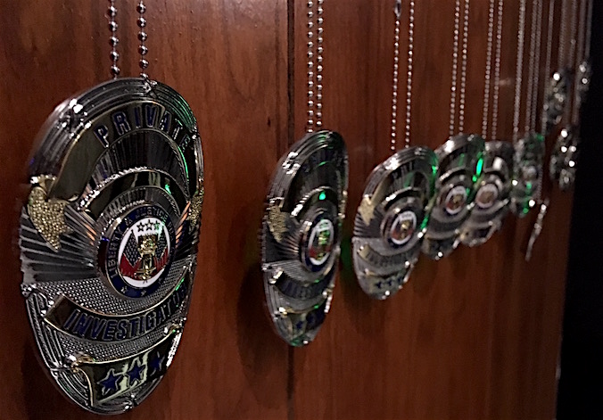 A line of a dozen "Private Investigator" badges hanging on chains along a wood paneled wall. 