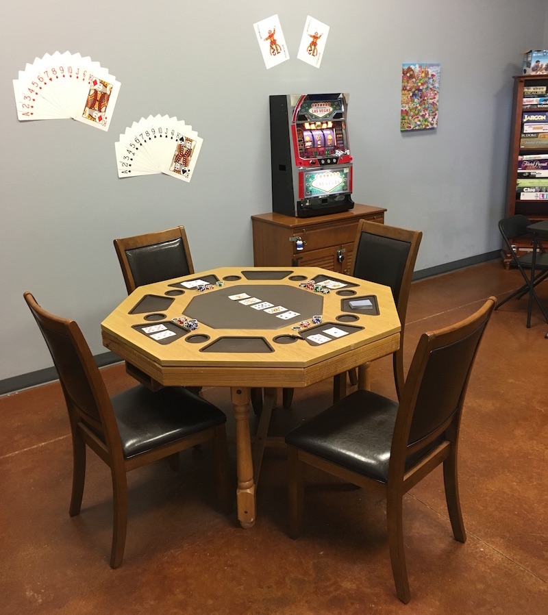 In game: a card table with a game of poker in progress sits in the foreground, assorted games and gaming related things reside in the background.