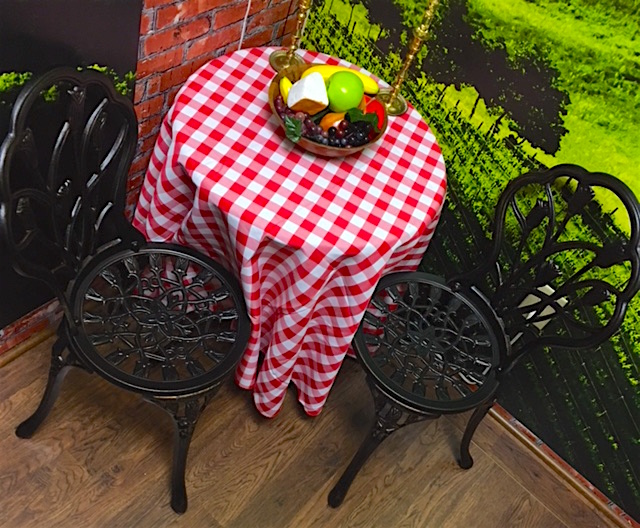 In-game: A table with a red & white checkered table cloth and two chairs. A bowl of fruit and cheese along with a pair of candle sticks rest atop the table.