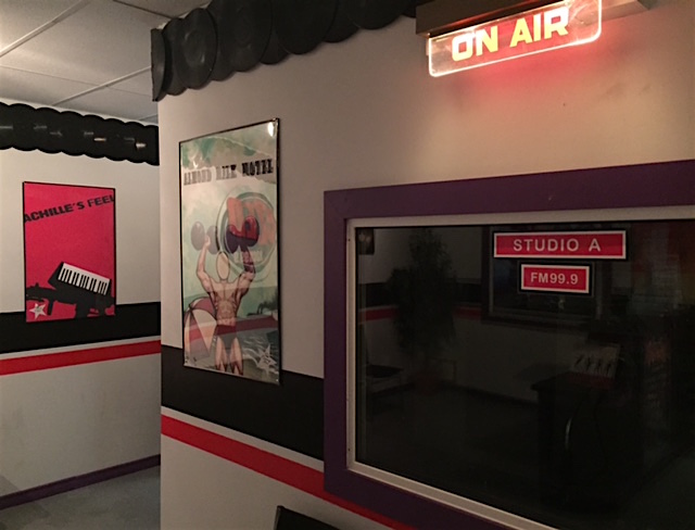 In-game: A studio with an "On Air" sign illuminated. Band art hangs on the walls.