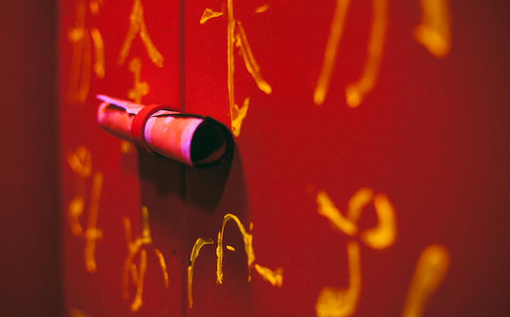 In-game: Close up of a wall with Japanese writing all over it and a small scroll rolled up and tied to the wall.