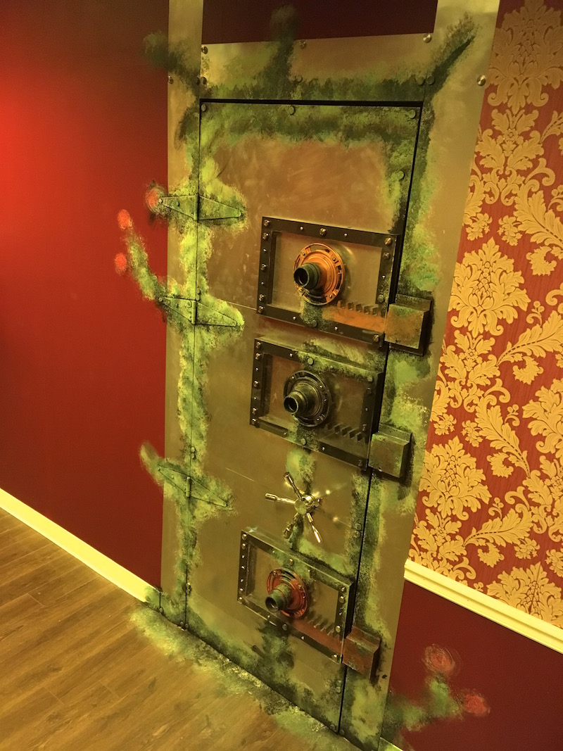In game: A big metal, vault door with a series of elaborate mechanical locking mechanisms and strange green weathering.