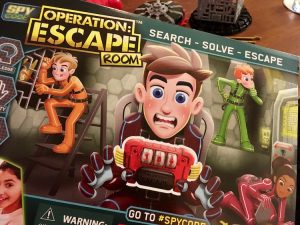 Spy Code Operation: Escape Room box features cartoon kids solving puzzles, and one kid in a chair sweating with the timer strap attached to him.