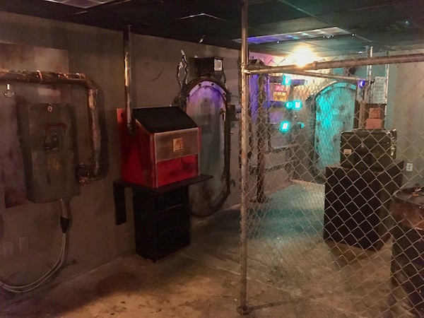 In-game: A wide angle image of the bunker. Doors and interactions lining the walls, a chainlink fence dividing part of the room. 