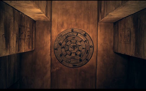 In-game: A narrow corridor in an Egyptian tomb with a circular series of wheels with symbols on the far wall.