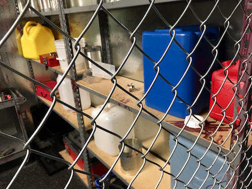 In-game: Shot through a gate, behind the wire is warehouse shelving with a variety of supplies.