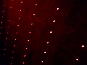 In-game: a dark room's wall covered in small, red, glowing lights.