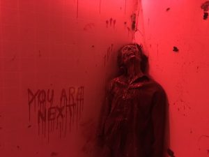 In-game: a dead body hung by a hook in his mouth hands next to a bloody message, "You are next."