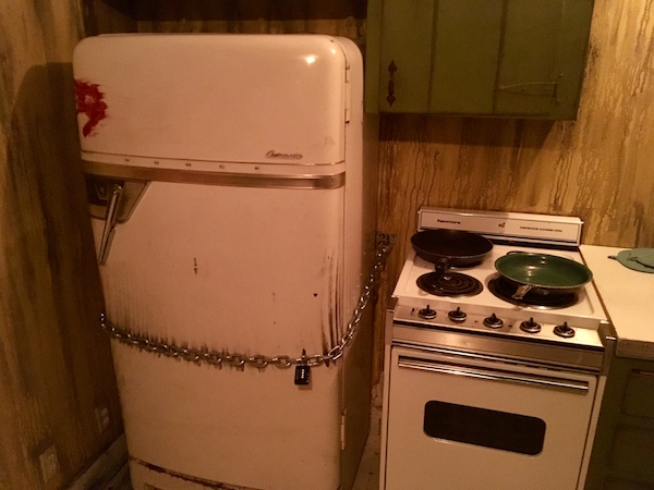 In-game: a rundown kitchen with an old stove and refrigerator locked shut with a chain and padlock.