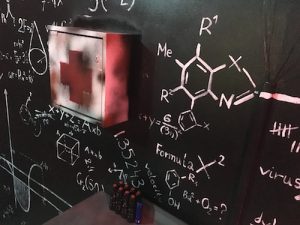 In-game: a blackboard covered in equations and molecules diagrams. There is a locked and worn first aid kit mounted to it.