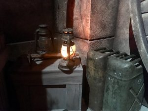 In-game: A close up of a lantern, water tanks, and a crate in front of an archeological dig site.