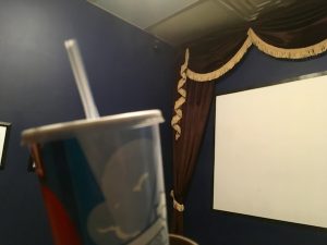 In-game: The screen of a movie theater in the background, a cup and straw in the foreground.