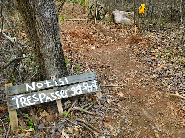 In-game: A hand painted wooden sign leans against a tree. It reads, "Notis! Trespasser get shot."