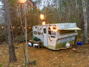 In-game: An old trailer in the middle of the woods. It's lit with a long strand of light bulbs.