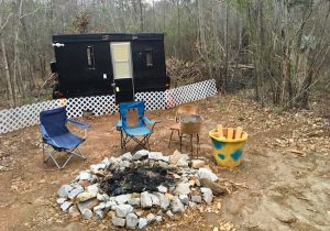 In-game: A trailer with a few folding chairs around an extinguished fire pit.