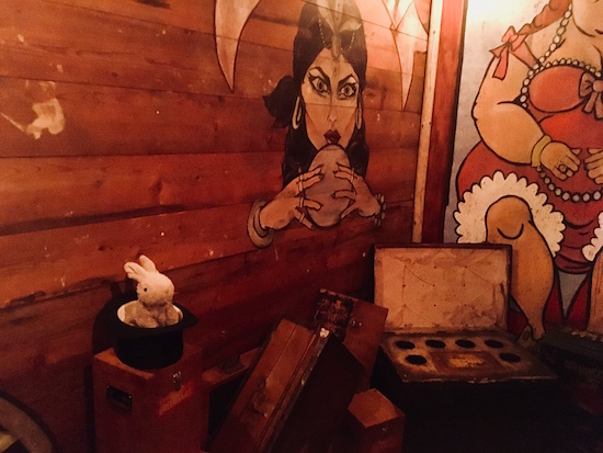 In-game: A series of boxes and props from the freakshow beside a wooden trailer. There is a rabbit in a top hat, and a the image of a fortune teller painted agains the wall.