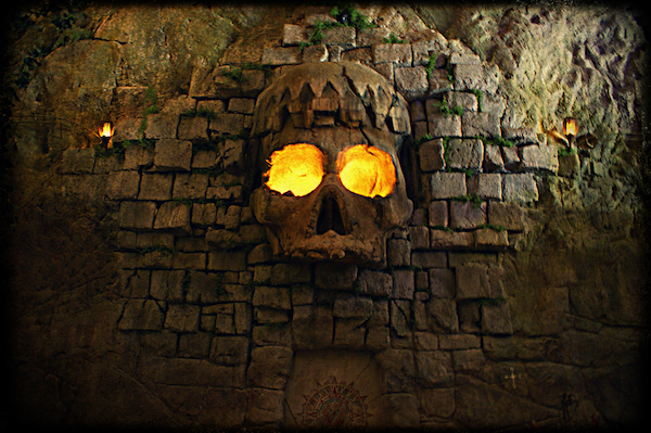 In-game: a large stone wall with a massive skull carved into it. The skull's eyes glow with fire.