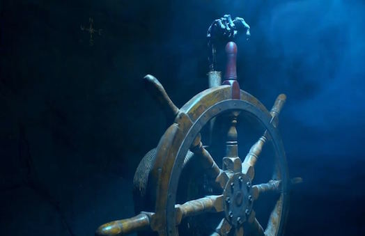 In-game: Wooden ship wheel with a skeletal hand pointing at the top of the wheel.
