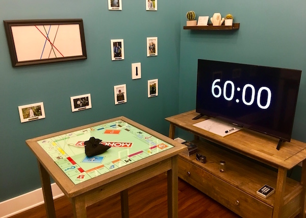In-game: Apartment A features a TV displays a 60 minute clock, a small table with a Monopoly board, and a wall of photos and cacti.