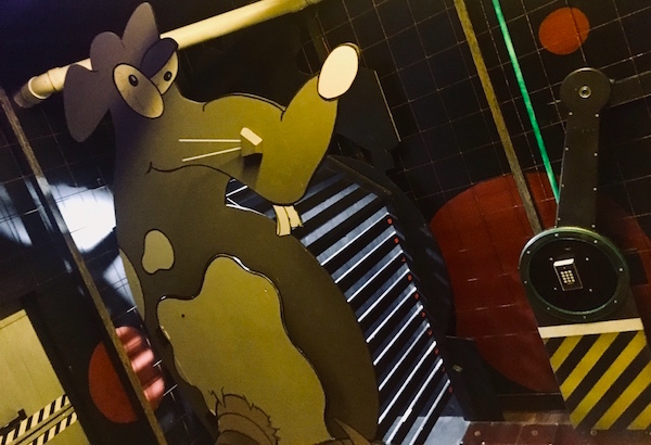 In-game: a dark rat cage with a large throw switch, a number pad, and an oversized caricature of a rat.