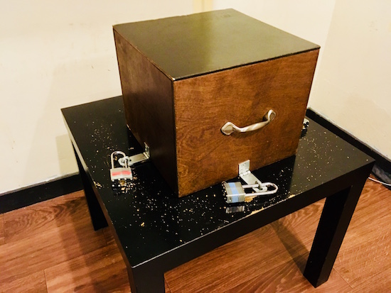 In-game: a wooden box locked down to a very beat up table by three padlocks.