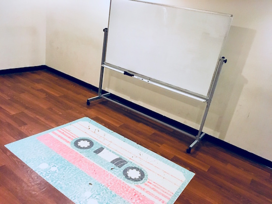 In-game: a plain white walled room with a whiteboard and a large fading cassette tape decal on the floor.