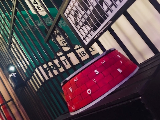 In-game: a food bowl with letters and symbols printed on it in a large cage. Beside it is a gigantic box of "Fruity Kibble."