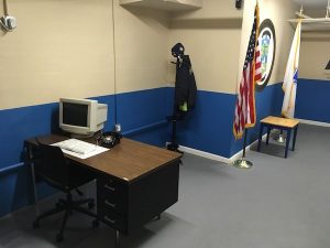 In-game: The police station, featuring an American & Massachusetts flags, a coat hanger with a uniform and a desk with a computer.