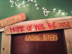 In-game: Christmas lights strung with the numbers 2 & 6 hanging from them. Below it, a door with wood panels that have the words, "Home of Will The Wise" and "Castle Byers," painted on them.