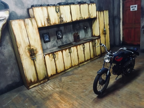 In-game: Wide angle shot of The Garage, a small motorcycle sits in the middle of the room, and a work bench and large set of cabinets sit in the background.