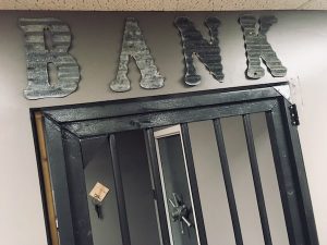 In-game: The word "BANK" cut from corrugated Aluminum. Below it is a barred door and beyond it, a vault door.