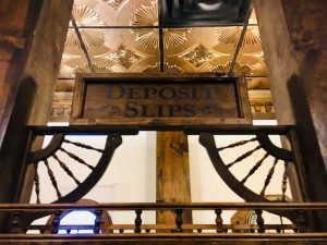 In-game: A sign that reads, "Deposit Slips" surrounded by intricate woodwork, above it is a beautiful gold ceiling.