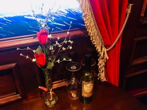 In-game: A vase with roses, two wine glasses, and a bottle on a table before a window with a snow storm beyond it.