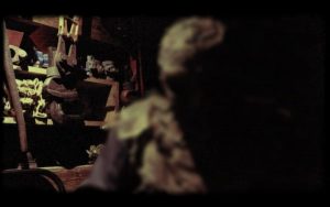 In-game: a menacing hooded man in a workshop.