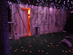 In-game: a cabin covered in ivy in the middle of the woods.