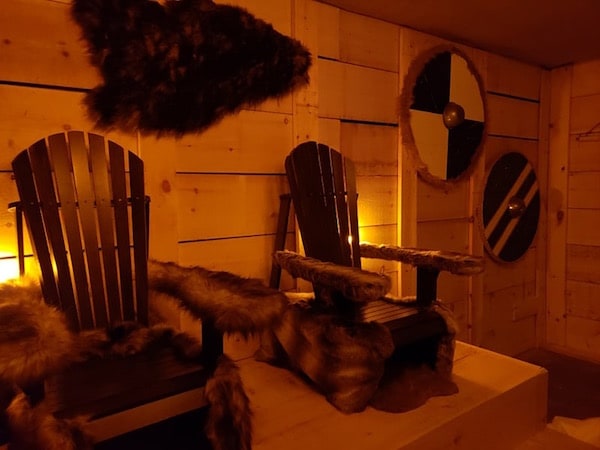In-game: The inside of a cabin with two wood and fur thrones surrounded by round shields.