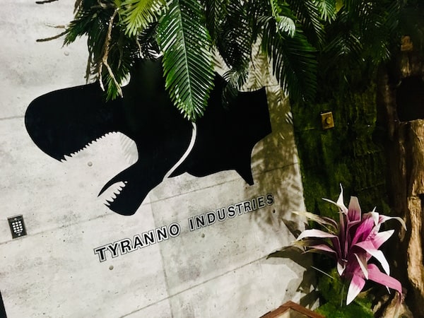 In-game: The Tryanno Industries logo on a concrete building in a tropical setting.
