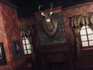 In-game: An elegant study with red and wood walls. The trophy of a buck hangs over a fireplace.