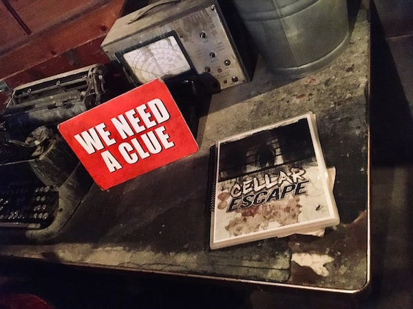In-game: A red we need a clue sign sitting beside a laminated and spiral-bound book labeled "CELLAR ESCAPE." All surrounded by a beautiful set.