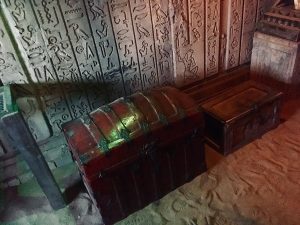 In-game: A treasure chest, and small locked wooden box sitting on top of sand with hieroglyphs adorning the wall behind them.