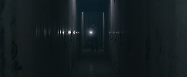 A man with a flashlight off in the distance down a long hall of storage units.