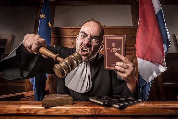 An enraged judge staring into directly into the camera, pointing with a gavel and holding up a book of Criminal Law.