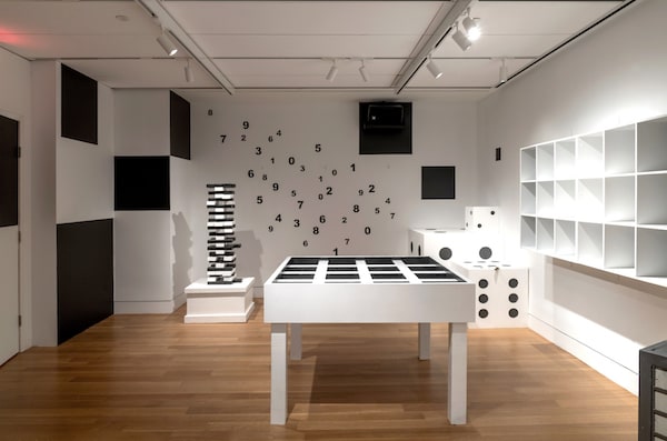 In-game: a black and white room with numbers on the wall, a large gridded table, oversized dice, and a tall jenga-like tower.