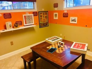 In-game: Wide shot of the game museum. The exhibits are laid out in a timeline, with classics such as Clue, Scrabble, and Bingo in view.