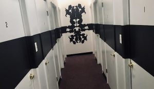 In-game: A long black and white hallway with doors on boths sides and a large rorschach image on the far wall.