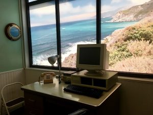 In-game: An old Tandy computre in front of a large window with a ocean view.