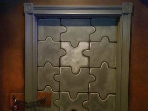 In-game: closeup of a gray door pained like a jigsaw puzzle and chained shut.
