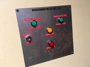In-game: Closeup of a communications panel with multiple lights and a big red button.